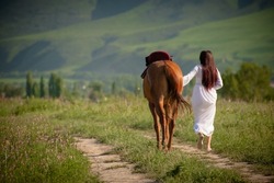 Asian girl (Kyrgyz) in a white dress walks with a horse in nature against the backdrop of mountains, Bishkek Kyrgyzstan
