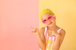 Girl swimmer in googles and swimming cap pointing with fingers aside at copy space on an pink studio background. Fashionable children's swimming goggles in the shape of a crab
