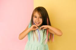 Cheerful girl with bright multicolored strands of hair laughs merrily looking at the cameras and straightening her hair with her hands. Child on a yellow-pink studio background. Stylish hair coloring
