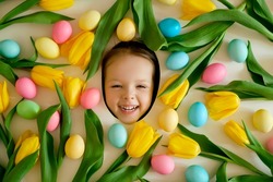 A child's face surrounded by tulip flowers and Easter painted multicolored eggs. Happy Easter holiday. The little handsome girl laughs funny with her mouth open. 