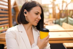 A thoughtful business woman in a white jacket with a disposable cup of tea looks away. A free moment to relax. Working Coffee Break. The person listens to music using wireless earphones