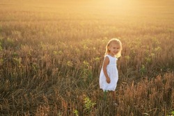 A little beautiful happy girl in a white linen dress walks in a mown wheat field at sunset on a hot summer evening. World Children's day