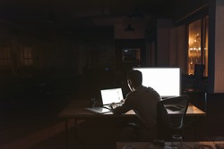 Rearview of a young businessman sitting alone at a desk in a dark office working on a laptop late in the evening