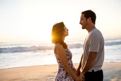 Loving young couple staring into each other's eyes while standing hand in hand together on a sandy beach at sunset
