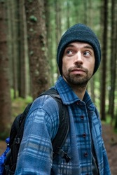 Young hiker in the woods. Sporty man with blue shirt and blue eyes he poses in a fir forest. Mountain holidays, endurance and high altitude sports. Portrait of handsome man with backpack and cap.