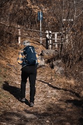 Elderly hiker walks along a mountain path. Back view of an old man with backpack and hiking boots. Peace, nature walk and healthy lifestyle. Gray hair and plaid shirt.