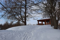 Beautiful winter landscape. A snowy path on the hillside, a wooden gazebo on the top. Snowdrifts, trees with a spreading crown. The ground is covered with snow. Blue sky and clouds. Picturesque nature