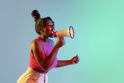 Profile picture of young beautiful African girl, female model speaking into megaphone isolated on blue green background in neon light. Concept of human emotion, facial expression. Copy space for ad.