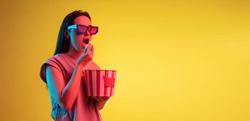 Excited. Watching cinema. Young pretty caucasian girl in 3d glasses with bucket of popcorn isolated on yellow studio background in neon light. Flyer. Copyspace for ad.
