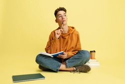 Pensive, thoughtful. One young caucasian guy, student in glasses sits on floor with notepad and books on yellow studio background. Education, studying and student life concept.