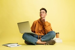 Studying, doing homework. One young smiling caucasian man, student in glasses sits on floor with laptop isolated on yellow studio background. Education, studying and student life concept.