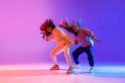 Active lifestyle. Two beautiful hip-hop dancers in motion on gradient pink purple neon background. Sport achievement, expression. Concept of dance, youth, hobby, dynamics, movement, action, ad