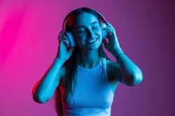 Close-up portrait of young beautiful girl wearing headphones and listening music on gradient pink purple background. Creative lifestyle, art importance. Concept of youth culture, music, art, ad
