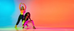 Portrait of two young beautiful hip-hop female dancers in modern clothes on colorful gradient blue orange at dance hall in neon. Youth culture, movement, active lifestyle, action, street dance, ad