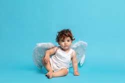 Happy childhood. Little angel. Portrait of cute toddler boy, baby in white romper with angel wings isolated on blue studio background. Concept of childhood, motherhood, life, birth. Copy space for ad