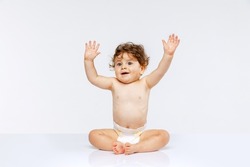 Portrait of beautiful playful toddler boy, baby in diaper sitting with raised hands isolated over white studio background. Concept of childhood, motherhood, life, birth. Copy space for ad