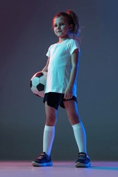 Portrait of little girl, football player standing with ball, posing isolated on gradient blue background in neon. Concept of action, sportive lifestyle, team game, health, education, childhood and ad