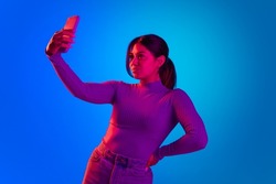 Young beautiful girl taking selfie with phone front camera isolated over blue background in pink neon light. Concept of beauty, youth, facial expression, emotions, lifestyle. Copy space for ad