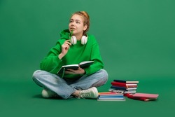 Portrait of young girl, student in casual cloth sitting on floor with thoughtful expression, studying isolated over green studio background. Concept of education, studying, homework, youth, lifestyle