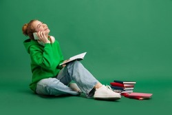 Portrait of young girl, student in casual cloth, sitting on floor, cheerfully talking on phone isolated over green studio background. Concept of education, studying, homework, youth, lifestyle