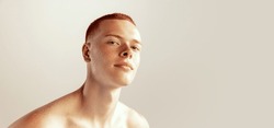 Portrait of handsome young red-haired man with freckles posing, barely smiling, looking at camera isolated over grey studio background. Concept of men's health, lifestyle, beauty, body and skin care