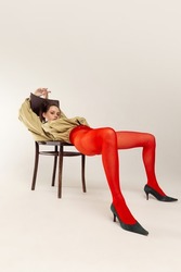 Portrait of stylish young girl in red tights and jacket posing, sitting on chair isolated over grey studio background. Concept of retro fashion, art photography, style, queer, beauty