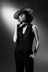 Black and white photography of stylish, elegant woman in classical black suit and stylish hat smoking, posing. Coco Chanel style. Monochrome effect. Concept of fashion, style, history