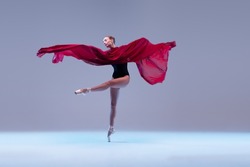 Portrait of young ballerina dancing with deep red clored fabric isolated over blue grey studio background. Tiptoe movements. Concept of classic ballet, inspiration, beauty, dance, creativity
