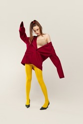 Portrait of young stylish girl in yellow tights and red jacket posing isolated over grey studio background. Freedom of style. Concept of retro fashion, art photography, style, queer, beauty