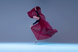 Portrait of young flexible ballerina dancing with transparent fabric isolated over blue grey studio background. Deep red cloth. Concept of classic ballet, inspiration, beauty, dance, creativity