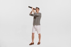 Portrait of young man, sailor in striped shirt looking in spyglass isolated on white background. Curious person. Concept of summer holidays, occupation, retro fashion, vintage style. Copy space for ad