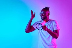Angry african-american man in glasses shouting in megaphone and holding one hand up over gradient colorful background in neon. Concept of people, emotions, expression, job, ad