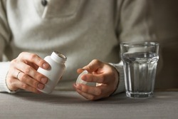 Close-up man's hands holds white plastic bottle with pills and ready to take supplement pill with glass of water. Ill man taking medication. Sick person need medicine against a headache and cold.
