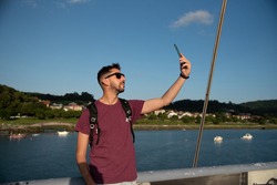 Latín Man with sunglasses taking photos with his mobile phone with a Maroon t-shirt on a sunny day with a clear blue Sky and a River with boats at his back