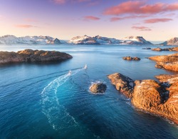 Aerial view of fishing boats, rocks in the blue sea, snowy mountains and colorful purple sky with red clouds at sunset in winter in Lofoten islands, Norway, Landscape with two ship. Top view. Travel