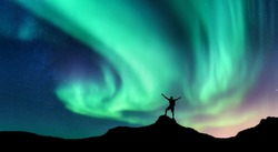 Aurora and silhouette of standing man with raised up arms on the mountain in Norway. Aurora borealis and happy man. Starry sky, green polar lights. Night landscape. Northern lights. Travel and tourism