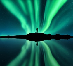 Aurora borealis and silhouette of standing man. Lofoten islands, Norway. Aurora and happy man. Stars and green polar lights. Night landscape with aurora, man, lake, sky reflection in water. Travel