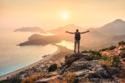 Happy sporty man with backpack and raised up arms standing and on the rock and looking at the seashore and mountains at sunset in summer. Man, sea, mountain ridges and orange sky. Oludeniz, Turkey