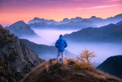 Sporty man on the mountain peak looking on mountain valley with low clouds at colorful sunset in autumn in Dolomites. Landscape with traveler, foggy hills, forest in fall, amazing sky at dusk in Alps