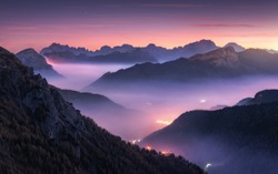 Mountains in fog at beautiful night in autumn in Dolomites, Italy. Landscape with alpine mountain valley, low clouds, forest, purple sky with stars, city illumination at sunset. Aerial. Passo Giau