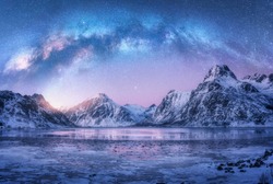 Milky Way above frozen sea coast and snow covered mountains in winter at night in Lofoten Islands, Norway. Arctic landscape with blue starry sky,  water, ice, snowy rocks, milky way. Space and galaxy