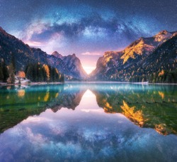 Milky Way reflected in water in mountain lake at starry night. Autumn landscape with purple sky with stars, blue fog, reflection, trees with colorful leaves, rocks in fall at sunset. Space and galaxy