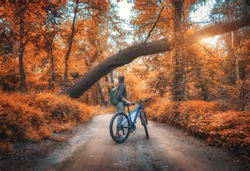 Woman riding a bicycle in  forest in autumn at sunset. Colorful landscape with sporty girl with backpack riding a mountain bike, dirt road, trees with orange leaves  in fall. Sport and travel. Cycle