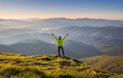 Sporty man standing on the mountain peak with raised up arms against mountain valley in fog at sunset in autumn. Happy young man, rocks, forest and blue sky in fall. Traveler hiking in mountains