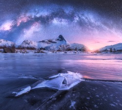 Milky Way above frozen sea coast and snow covered mountains in winter at night in Lofoten Islands, Norway. Arctic landscape with blue starry sky,  water, ice, snowy rocks, milky way. Space and galaxy