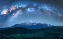Arched Milky Way over the beautiful mountains with snow covered peak at night in summer. Colorful landscape with bright starry sky with Milky Way arch, snowy rocks, hills. Galaxy. Nature and space