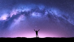 Arched Milky Way and happy man at night. Silhouette of guy with raised up arm on the hill, purple sky with stars, pink light in summer. Galaxy. Space background. Landscape with milky way arch. Travel