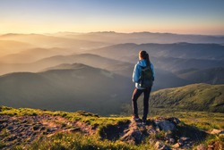 Girl on mountain peak with green grass looking at beautiful mountain valley in fog at sunset in summer. Landscape with sporty young woman, foggy hills, forest, sky. Travel and tourism. Hiking