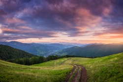 Mountain dirt road at beautiful sunset in summer. Colorful landscape with road, meadows with green grass, sky with vibrant clouds, mountains with forest. Trail on the hill. Travel and nature. Scenery
