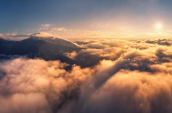 Mountains in clouds at sunrise in summer. Aerial view of mountain peak in fog. Beautiful landscape with high rocks, forest, sky. Top view from drone of mountain valley in low clouds. Foggy hills
